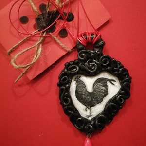 Op de afbeelding: Een zwarte hangerketting met een hartvormig frame met een hanenillustratie. De hanger hangt aan een rood koord en heeft een roze druppelvormige kraal. Een rode kaart met touw en zwarte waszegels staat op de achtergrond.