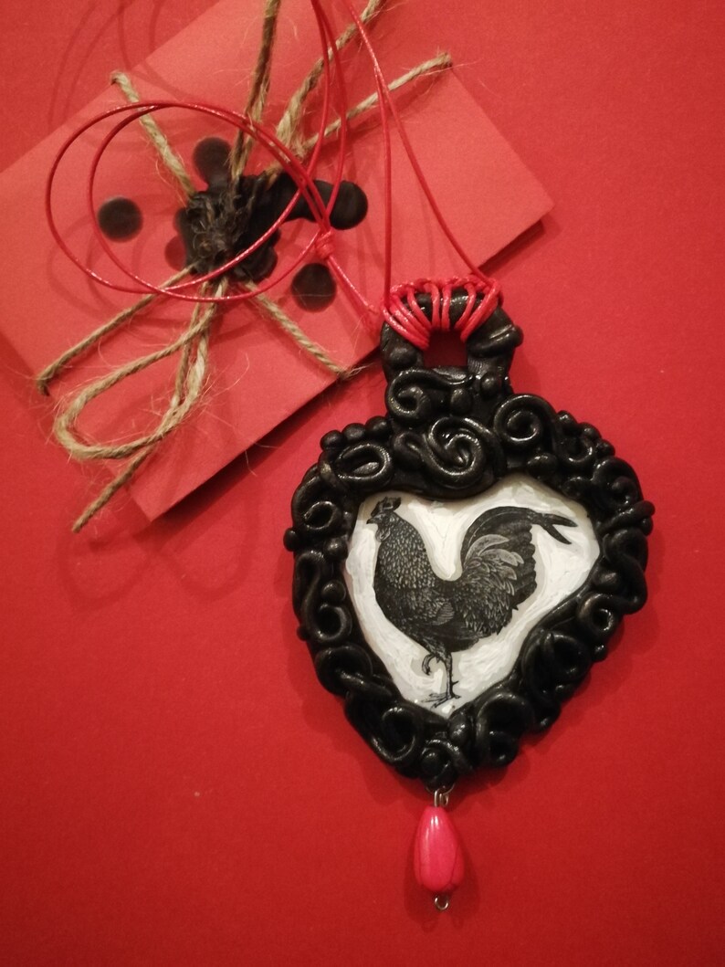 Op de afbeelding: Een zwarte hangerketting met een hartvormig frame met een hanenillustratie. De hanger hangt aan een rood koord en heeft een roze druppelvormige kraal. Een rode kaart met touw en zwarte waszegels staat op de achtergrond.