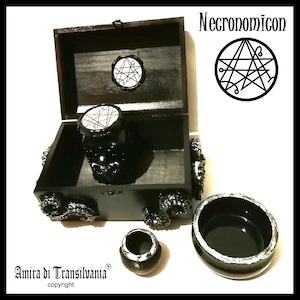 May include: A black wooden box with a hinged lid, containing a black skull-shaped jar and two small black containers. The box and jar are decorated with white accents. The word "Necronomicon" is displayed above a circular symbol.