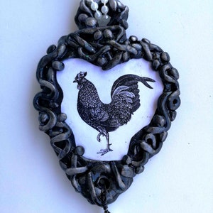 Op de afbeelding: Een hartvormige hanger met een zwart en zilveren frame, met een zwart-witte illustratie van een haan. De hanger is versierd met een witte parel en een snoer van witte kralen.