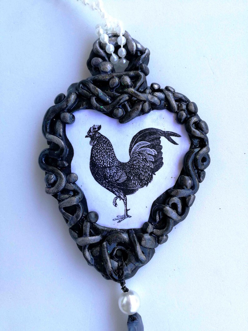 Op de afbeelding: Een hartvormige hanger met een zwart en zilveren frame, met een zwart-witte illustratie van een haan. De hanger is versierd met een witte parel en een snoer van witte kralen.