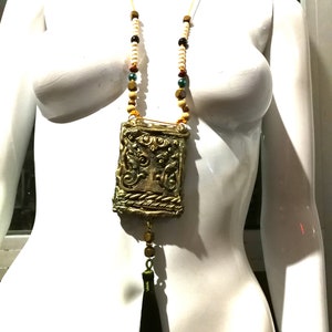May include: A gold necklace with a decorative pendant and a black tassel. The pendant is a rectangular box with intricate designs and a rope-like border. The necklace is made of wooden beads in various colors, including beige, brown, red, and green.