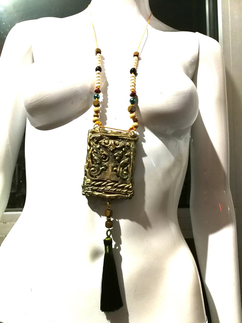 May include: A gold necklace with a decorative pendant and a black tassel. The pendant is a rectangular box with intricate designs and a rope-like border. The necklace is made of wooden beads in various colors, including beige, brown, red, and green.