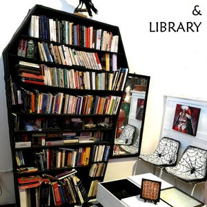 May include: A black coffin-shaped bookshelf filled with books, topped with a black crow statue. The text "AMIRA MAGIC STUDIOS & LIBRARY" is displayed above the bookshelf. Two chairs with spiderweb designs and a white coffee table are also visible.