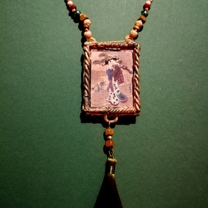 May include: A gold-toned pendant necklace with a framed image of a woman in a traditional Japanese kimono. The necklace features a black tassel and a beaded chain with white, brown, and green beads.