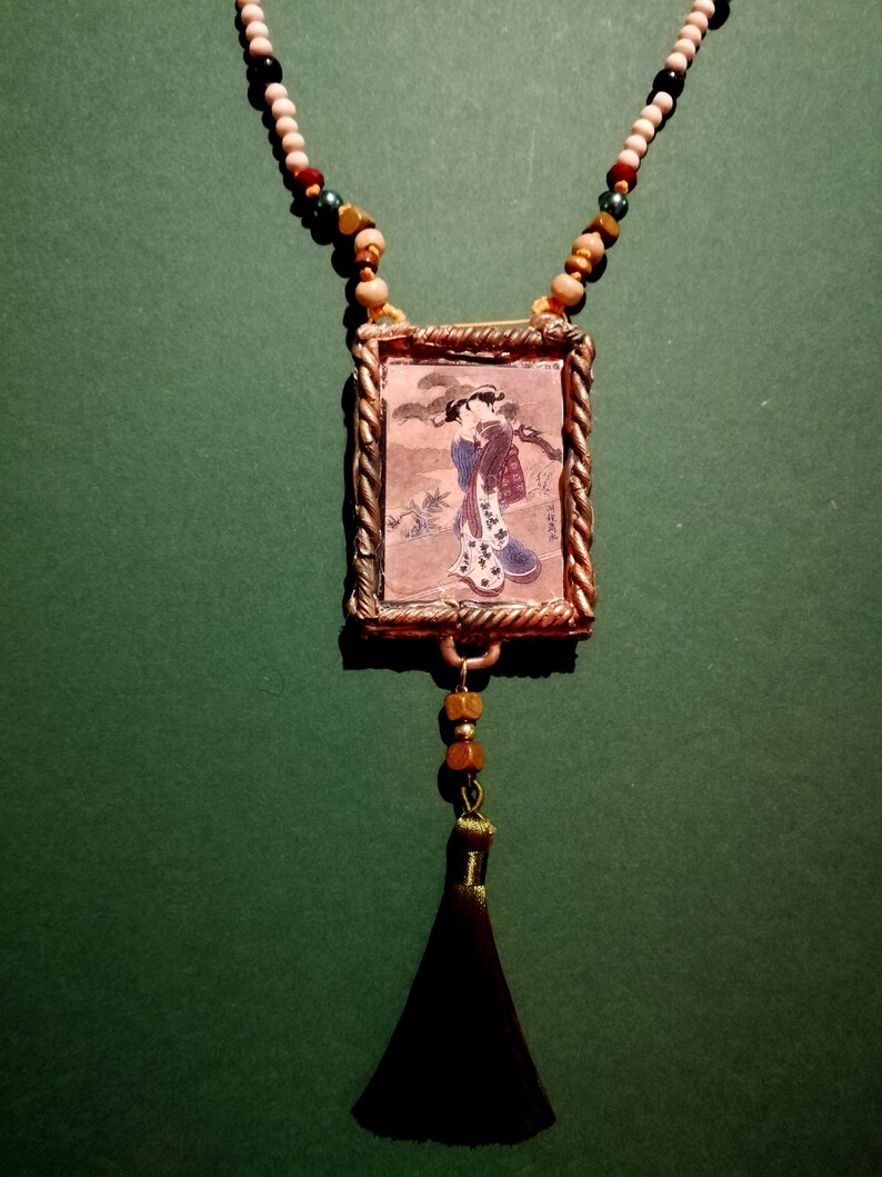 May include: A gold-toned pendant necklace with a framed image of a woman in a traditional Japanese kimono. The necklace features a black tassel and a beaded chain with white, brown, and green beads.
