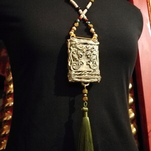 May include: A silver pendant necklace with a detailed ornate design, featuring a tassel and a variety of beads in white, brown, gold, and green.