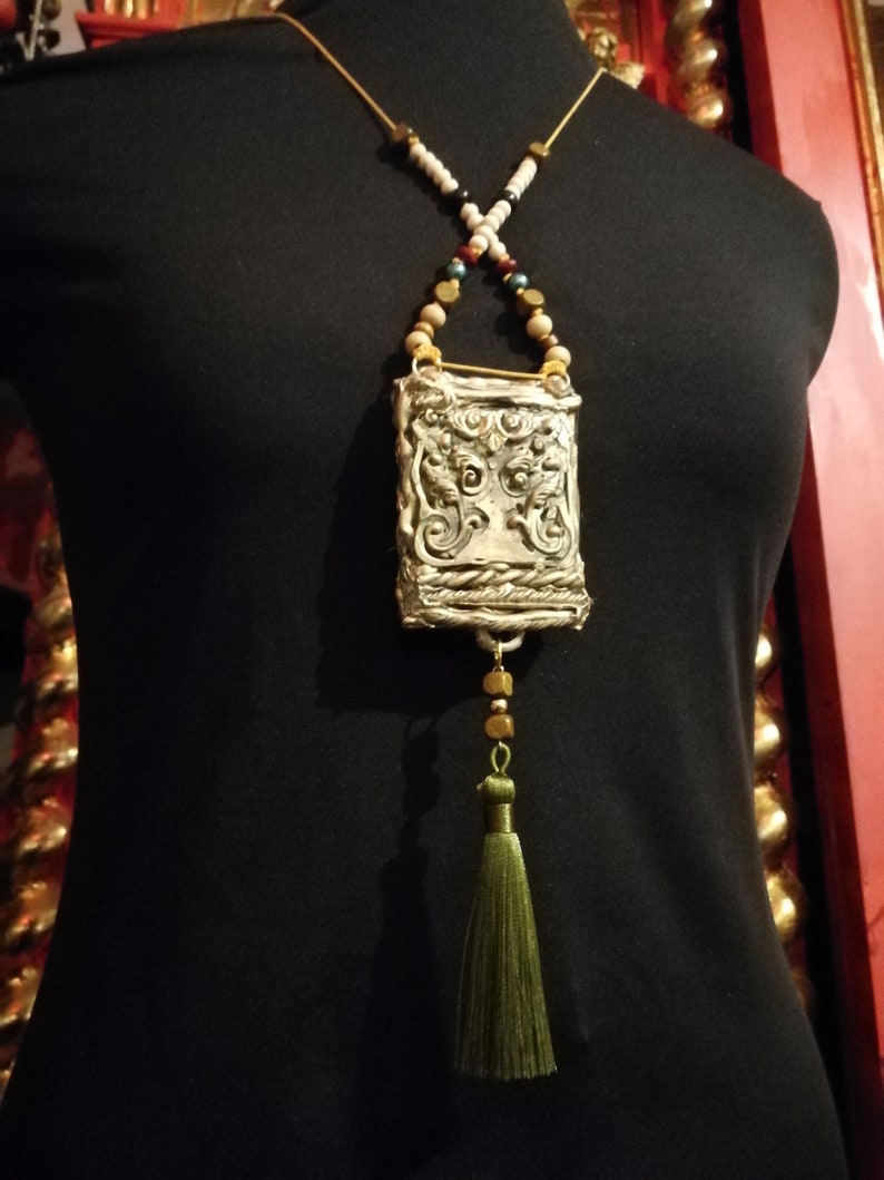 May include: A silver pendant necklace with a detailed ornate design, featuring a tassel and a variety of beads in white, brown, gold, and green.