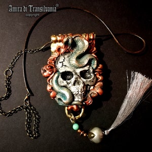 Puede incluir: Un cráneo plateado con una serpiente verde envuelta alrededor, colocado en un marco de cobre y oro. El cráneo es parte de un collar con una cadena plateada, una cuenta turquesa, una cuenta marrón, una cuenta plateada y un borla plateada.
