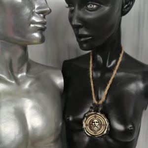 May include: A black and silver mannequin bust wearing a gold pendant necklace with a circular design featuring a face. The pendant is on a brown rope chain.