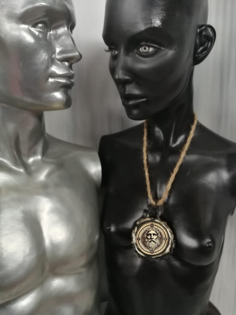 May include: A black and silver mannequin bust wearing a gold pendant necklace with a circular design featuring a face. The pendant is on a brown rope chain.