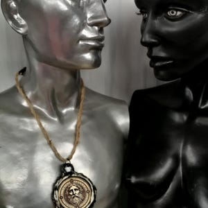 May include: A silver and black mannequin wearing a necklace with a round pendant featuring a carved face. The pendant is made of metal and has a brown rope cord.