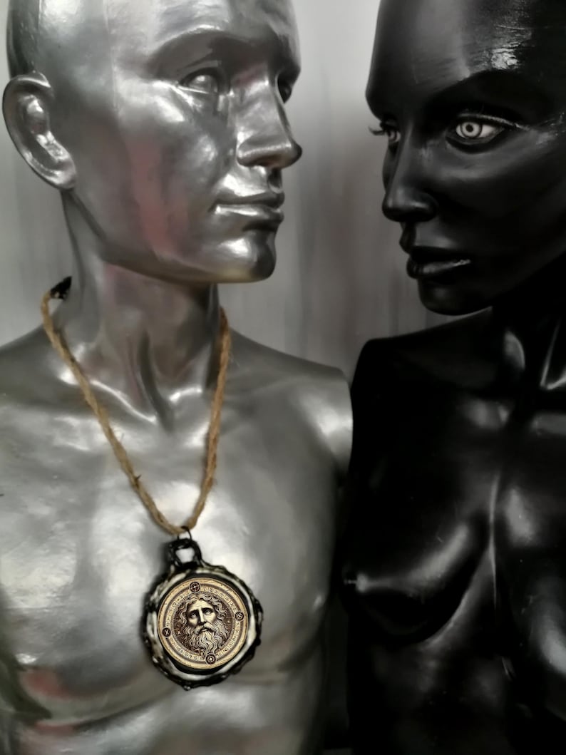 May include: A silver and black mannequin wearing a necklace with a round pendant featuring a carved face. The pendant is made of metal and has a brown rope cord.