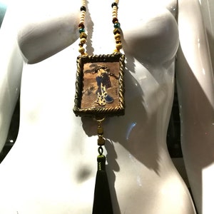 May include: A gold framed pendant necklace with a black tassel. The pendant features a vintage illustration of a woman in a kimono. The necklace is made with white, brown, and green beads.