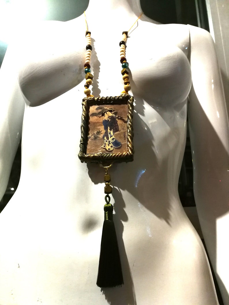 May include: A gold framed pendant necklace with a black tassel. The pendant features a vintage illustration of a woman in a kimono. The necklace is made with white, brown, and green beads.