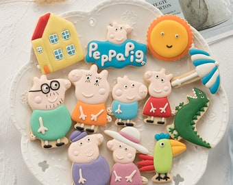 May include: A plate of decorated sugar cookies featuring characters from the children's cartoon show Peppa Pig. The cookies are decorated with colorful icing and include Peppa Pig, George Pig, Daddy Pig, Mummy Pig, and other characters. The cookies are arranged on a white plate with a scalloped edge.