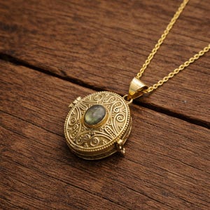 Könnte beinhalten: Eine goldfarbene Medaillon-Halskette mit einem ovalen Edelstein. Das Medaillon hat aufwendige, geprägte Details und eine zarte Kette. Der Edelstein ist von gedeckter grüner Farbe. Die Halskette liegt auf einer Holzoberfläche.