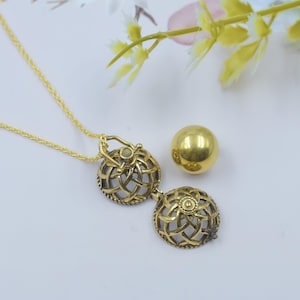 Puede incluir: Un collar dorado con un colgante con un diseño calado. El colgante es esférico y se abre para revelar un pequeño compartimento. También hay una esfera dorada separada, lo que sugiere un conjunto o un componente adicional.