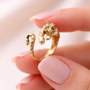 May include: A gold-colored ring in the shape of a tiger. The ring features a detailed tiger design with the tiger's head and paws. The ring is held by a person with pink nail polish.