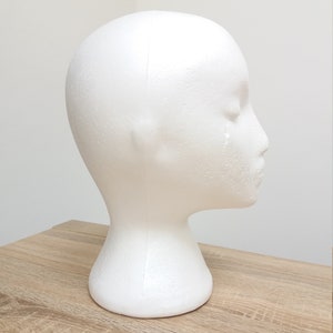 May include: White foam mannequin head for displaying wigs, hats, or other accessories. The head is on a wooden surface.
