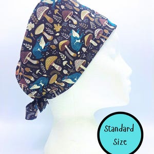 May include: A dark blue surgical scrub cap with a whimsical pattern of mushrooms, frogs, and foliage. The cap is tied at the back and is displayed on a mannequin head. The text "Standard Size" is in a blue circle.