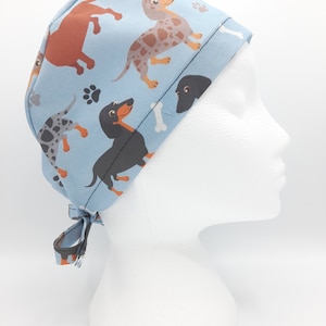 May include: A blue surgical scrub hat with a pattern of dachshunds in various colors and poses. The hat has a tie at the back.