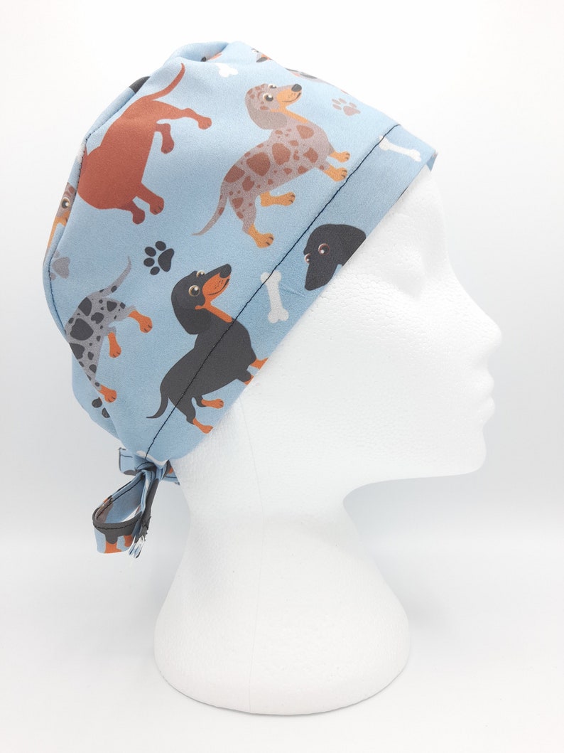 May include: A blue surgical scrub hat with a pattern of dachshunds in various colors and poses. The hat has a tie at the back.