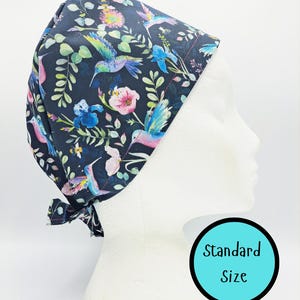 May include: A dark navy blue scrub cap featuring a colorful pattern of hummingbirds, bees, and various flowers in shades of pink, blue, and green. The cap includes a tie-back closure and a circular graphic with the text Standard Size.