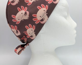 May include: A brown scrub hat with a pink and red axolotl pattern. The hat has a tie at the back.