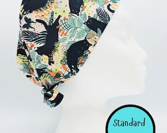 May include: A patterned scrub cap featuring black cats among green leaves and orange flowers on a white background. The cap is tied at the back. The text "Standard Size" is in a teal circle.