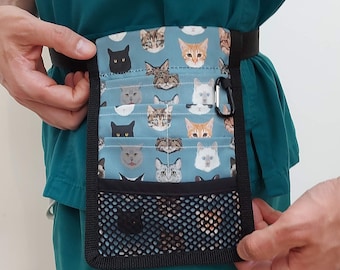 May include: A blue and black cat-themed utility belt with multiple pockets and a mesh pocket. The belt is worn by a person with a teal scrub top.