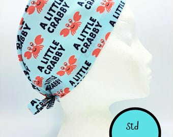 May include: A light blue scrub cap featuring a repeating pattern of orange cartoon crabs and the black text A LITTLE CRABBY. The cap has a tie-back closure and is displayed on a white mannequin head. A circular graphic indicates Std Size.