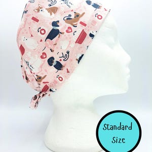 May include: A pink scrub cap featuring a pattern of dogs, cats, hearts, and medical items. The cap is tied at the back and displayed on a white mannequin head. The text "Standard Size" is in a blue circle.