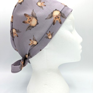 Puede incluir: Un gorro de quirófano morado con un patrón de murciélagos marrones. El gorro tiene un lazo en la parte posterior.