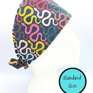 May include: A navy blue scrub cap with a vibrant snake design in red, yellow, pink, and white. The cap is tied at the back and is displayed on a white mannequin head. The text "Standard Size" is in a blue circle.