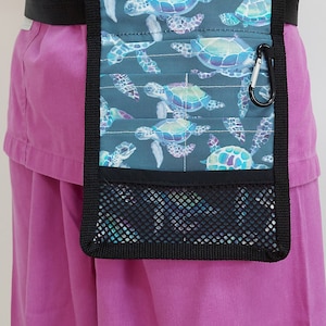 Könnte beinhalten: Ein schwarzer Stoffgürtel mit einer Tasche in Teal- und Lilatönen mit einem See-Schildkröten-Muster. Die Tasche hat ein Netzfach und einen schwarzen Karabinerhaken.