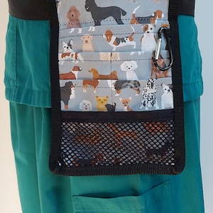 May include: A teal green scrub top with a black fabric belt and a black and grey dog-themed utility belt with a mesh pocket. The utility belt has a black carabiner clip.