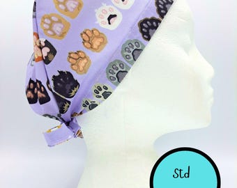 May include: A light purple scrub cap featuring a repeating pattern of multi-coloured animal paw prints in shades of orange, white, grey, and black. The fabric cap has a tie-back closure and a circular graphic with the text Std Size.