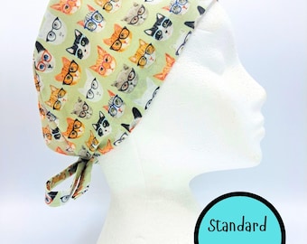 May include: A light green scrub cap featuring a repeating pattern of various cats wearing colourful eyeglasses. The fabric cap has a tie-back closure and includes a circular graphic with the text Standard Size in black lettering.