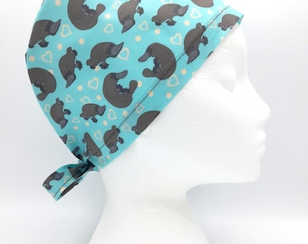 May include: A turquoise blue surgical scrub hat with a pattern of brown platypuses and white hearts. The hat has a tie at the back.