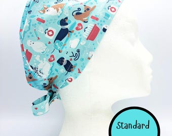 May include: A light blue surgical scrub cap featuring a pattern of dogs, cats, and medical items. The cap is tied at the back and displayed on a mannequin head. The text "Standard Size" is in a blue circle.