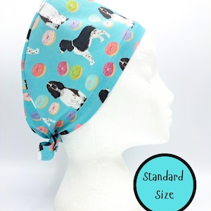 Puede incluir: Un gorro quirúrgico azul claro con un estampado de perros blancos y negros y donuts coloridos. El gorro está atado en la parte posterior y se muestra en una cabeza de maniquí blanca. El texto "Standard Size" está en un círculo azul.