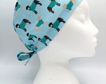 May include: A light blue surgical scrub hat with a pattern of dogs wearing surgical masks. The hat has a tie at the back.
