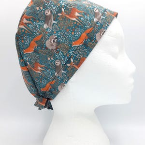 May include: A scrub cap featuring a teal background with a repeating pattern of woodland animals including orange foxes, brown deer, grey raccoons, and owls nestled among green foliage and small orange berries, tied at the back with fabric straps.