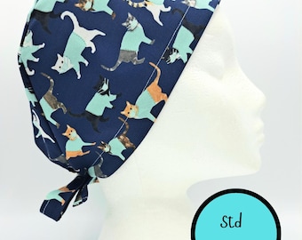 May include: A navy blue scrub cap featuring a repeating pattern of cats wearing light blue shirts and face masks. The cap has a tie-back closure and a circular graphic in the corner with the text Std Size in black lettering.