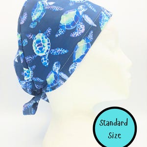 May include: A navy blue scrub cap featuring a pattern of sea turtles in shades of blue and green. The cap is tied at the back and is displayed on a mannequin head. The text "Standard Size" is in a teal circle.