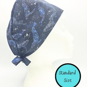 May include: A navy blue surgical scrub cap with a constellation design featuring whales and stars. The cap is tied at the back with a bow. The text "Standard Size" is in a teal circle.