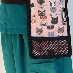 May include: A teal scrub top with a black belt and a pink and black cat-themed utility pouch with a mesh pocket. The pouch has a black carabiner clip.