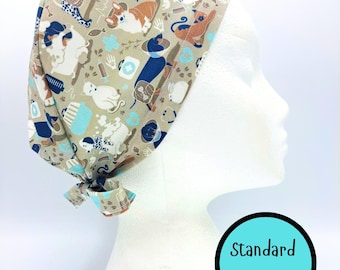 May include: A beige scrub cap featuring a repeating pattern of various dogs, cats, medical symbols, and veterinary equipment in shades of blue, white, and brown. The cap includes a tie-back closure and a circular graphic with the text Standard Size.