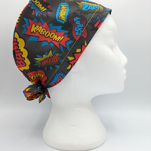 Op de afbeelding: Een zwarte scrub-muts met een stripboekprint met woorden als "Kaboom!" en "Zap!" in rood, geel en blauw.
